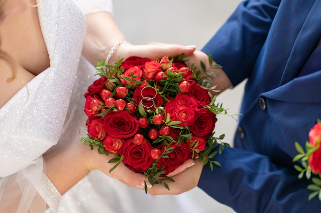 Die braut in einem weißen kleid und der bräutigam halten einen stilvollen hochzeitsstrauß aus roten rosen. hochzeitsdetails.