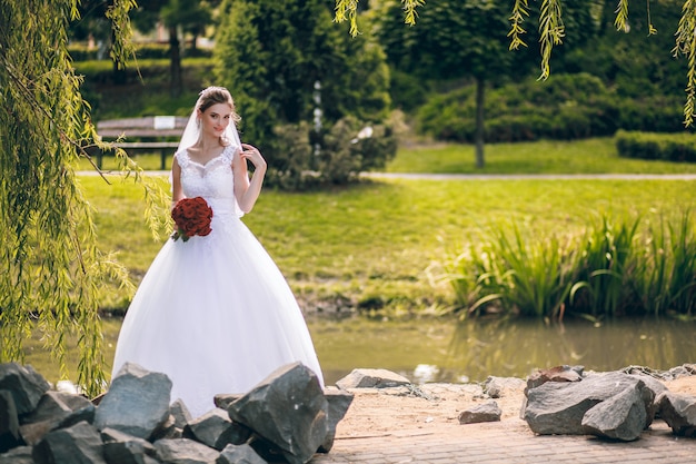 Die braut in einem weißen kleid und mit einem strauß roter rosen in den händen geht an einem warmen herbsttag in einem stadtpark spazieren. hochzeitsporträt.