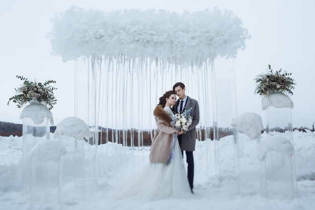 Die Braut mit einem schönen Blumenstrauß und der Bräutigam beim Hochzeitswinterschießen. Junge Leute stehen vor dem Hintergrund der Winterhochzeitsdekorationen