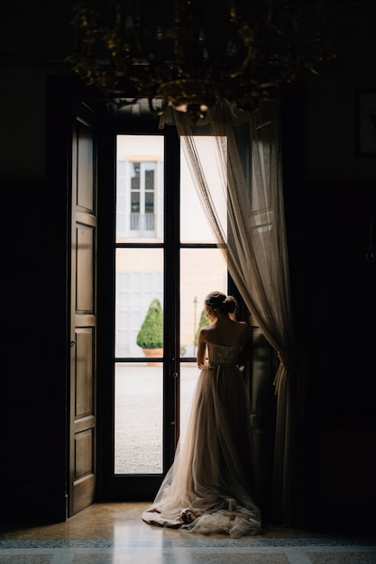 Die Braut steht mit gesenktem Kopf am großen Fenster einer alten Villa, Rückansicht