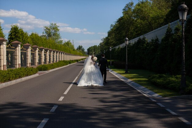 Die Braut und der Bräutigam gehen die asphaltierte Straße im Park entlang. Wind weht Brautschleier.