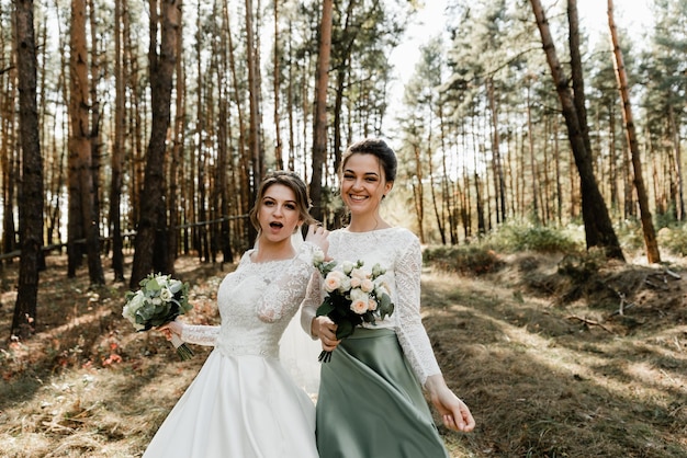 Die Braut und ihre Freundin in Hochzeitskleidern amüsieren sich im Wald und halten Blumensträuße in den Händen. weibliche Freundschaft. Hochzeitstag. Mädchen verwöhnen