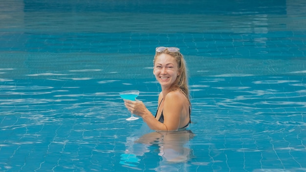 Die breite Mittelaufnahme hübsche Frau, die im Swimmingpool des Hotels blauen Cocktail-Alkohol trinkt Porträt eines sexy Mädchens im Pool im Freien