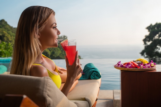 Die charmante blondine entspannt sich auf einer sonnenliege in der nähe des infinity-pools und trinkt einen wassermelonen-smoothie