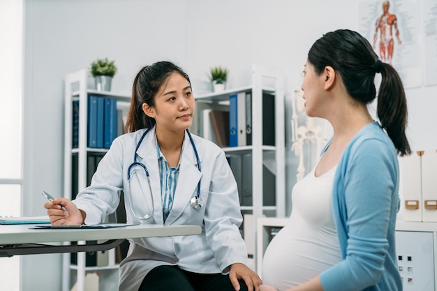 Die chinesische Gynäkologin, die einen Stift hält, hört aufmerksam zu, während ihre schwangere Patientin ihr von Problemen am Schreibtisch in einer modernen, hellen Klinik erzählt.