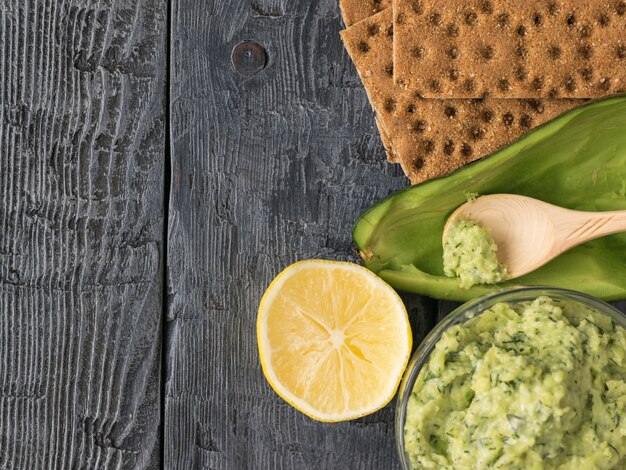 Die Creme der Kiwi in der Schüssel, im Toast, in der Zitrone und in den Kräutern auf einer Weinlesetabelle. Ansicht von oben. Von oben betrachten.