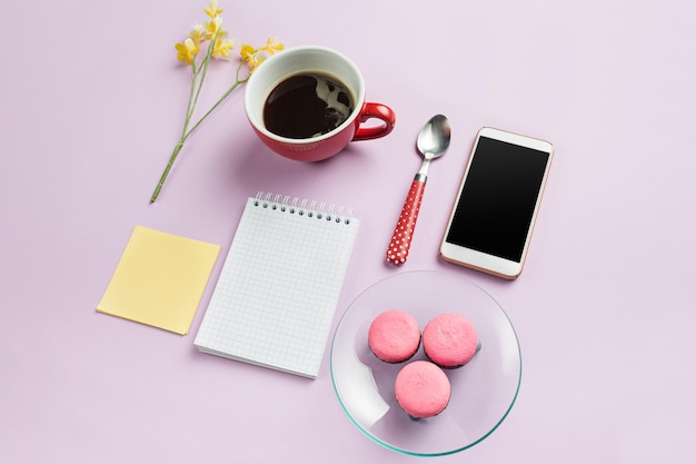 Die Draufsicht auf den weiblichen Schreibtisch. Mock-up mit französischen Macarons auf einem trendigen rosa Schreibtisch. Frau und stilvoller Arbeitsplatz. Tasse Kaffee, Telefon, Notizbuch. Frühstück, Telefon, Kaffee. Konzept zum Frauentag