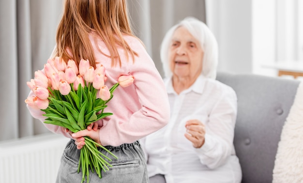 Die Enkelin im Vorschulalter überrascht die Großmutter und hält den Tulpenstrauß hinter ihren Rücken
