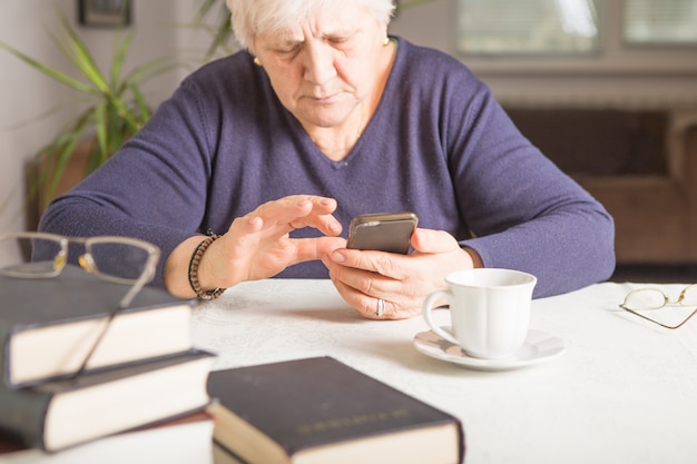 Die Frau im Ruhestand benutzt ein Mobiltelefon in der gemütlichen Küche zu Hause. Großmutter hat eine Kaffeepause beim Kochen