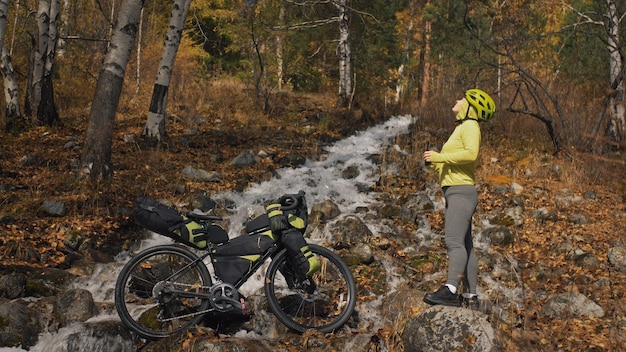 Die Frau reist auf gemischtem Gelände Radtouren mit Bikepacking Die Reise des Reisenden mit Fahrradtaschen Sportbekleidung in grün-schwarzen Farben Die Reise im magischen Herbstwald-Flussstrom