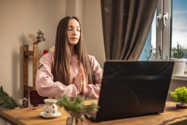 Die Frau sitzt an einem Tisch vor einem Laptop und meditiert und entspannt sich, während sie einem Ausbilder online oder einem Videovortrag zuhört Konzept einer Ferntherapie- und Meditationssitzung