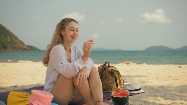 Die fröhliche Frau, die Wassermelonenscheiben auf einem tropischen Sandstrand hält und isst, porträtiert Attra