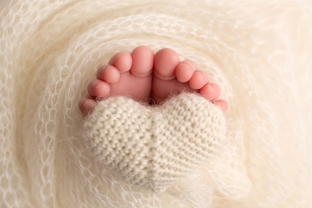 Die Füße eines Neugeborenen sind in eine gestrickte Decke gehüllt. Die Finger eines neugeborenen Babys halten ein weißes gestricktes Herz. Foto in hoher Qualität