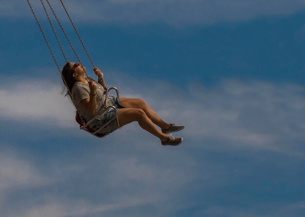 Die ganze Länge der jungen Frau, die auf einer Ketten-Swing-Fahrt gegen den Himmel schwingt