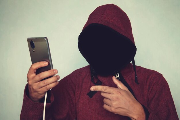 Die gesichtslose, nicht erkennbare Person mit Kapuze, die den Identitätsdiebstahl und das Technologiekriminalitätskonzept des Mobiltelefons verwendet, zeigt einen Finger auf einem Mobiltelefon