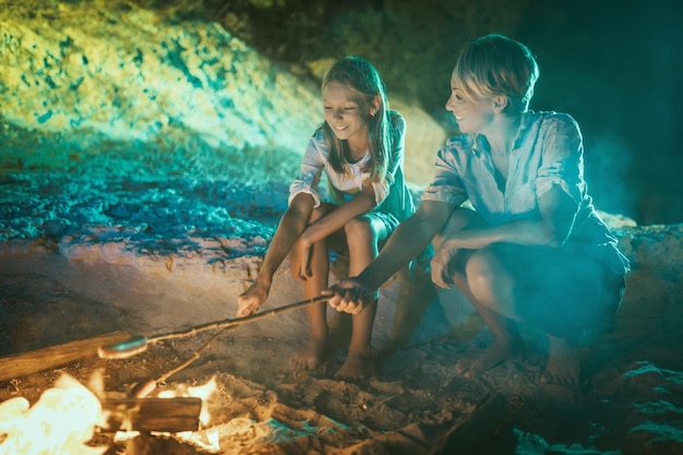 Die glücklich lächelnde Tochter und ihre Mutter sitzen abends am Feuer am sandigen Strand. Sie braten Würstchen am Stiel.