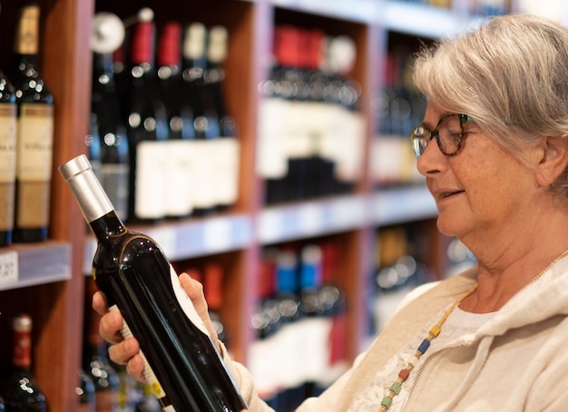 Die glückliche ältere Frau, die im Laden eine Flasche Rotwein wählt. Konsumkonzept