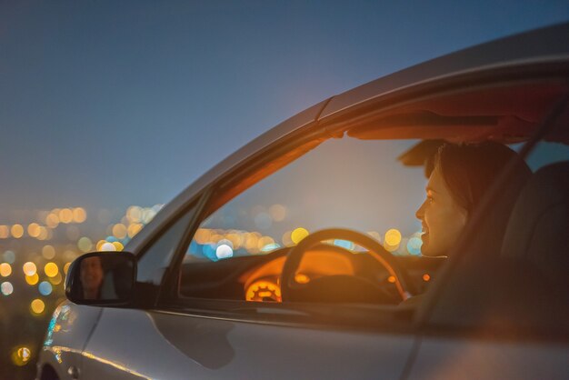 Die glückliche Frau sitzt in einem Auto auf dem hellen Hintergrund der Stadt. abend nacht zeit