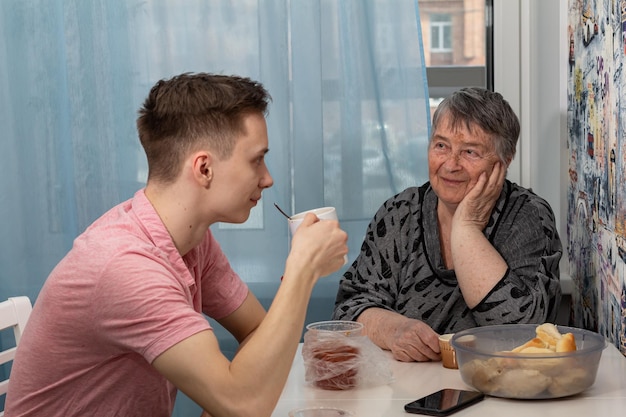 Die glückliche Großmutter und ihr erwachsener Enkel trinken Tee und diskutieren über die neuesten Nachrichten. Der Enkel mit selektivem Fokus unterstützt die Großmutter am globalen Tag der Eltern