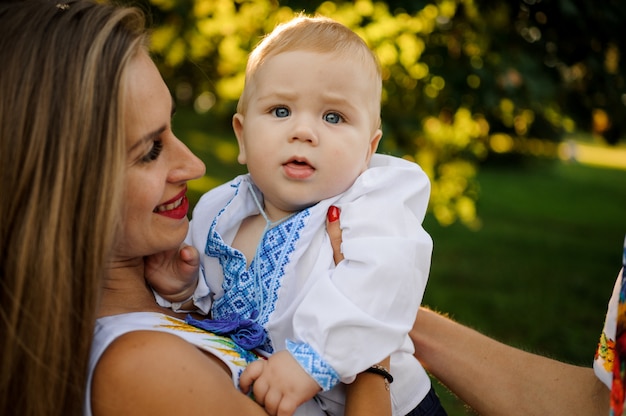 Die glückliche Mutter, die an Hände ein Baby hält, kleidete im gestickten Hemd an