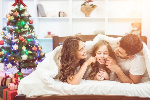 Die glückliche Tochter und die Eltern machen Spaß im Bett neben dem Weihnachtsbaum