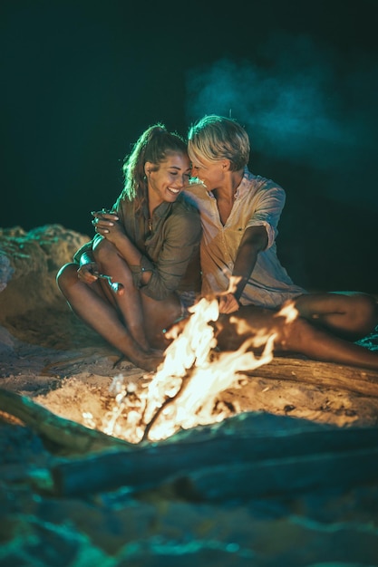 Die glücklichen zwei jungen lächelnden Frauen sitzen abends am Feuer am sandigen Strand. Sie braten Würstchen am Stiel.