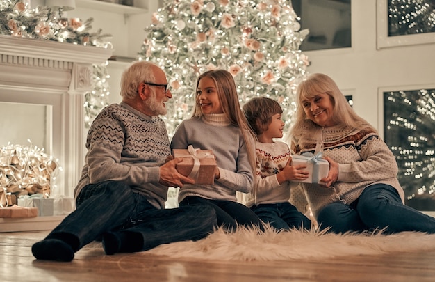 Die Großeltern, die Kindern auf dem Weihnachtsbaumhintergrund Geschenke geben