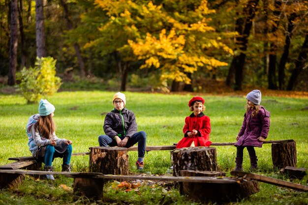 Die Gruppe kleiner Kinder, die sich im Herbstpark unterhalten Kinder in leichten Jacken sitzen auf einer provisorischen Holzbank