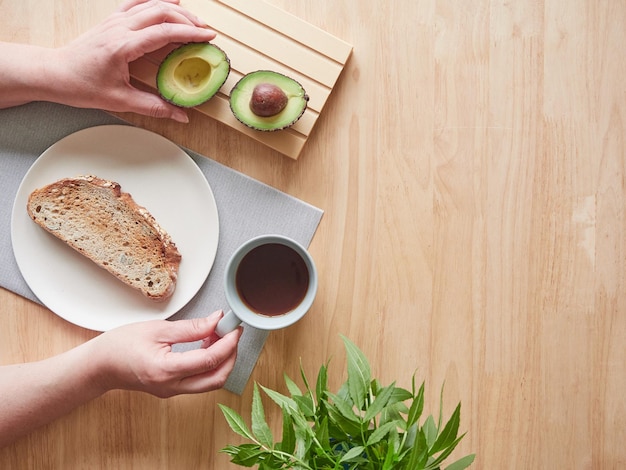 Die Hände der Frau, die Kaffeetasse halten und halbe Avocado auf Frühstückstisch aufheben Draufsicht mit Kopienraum