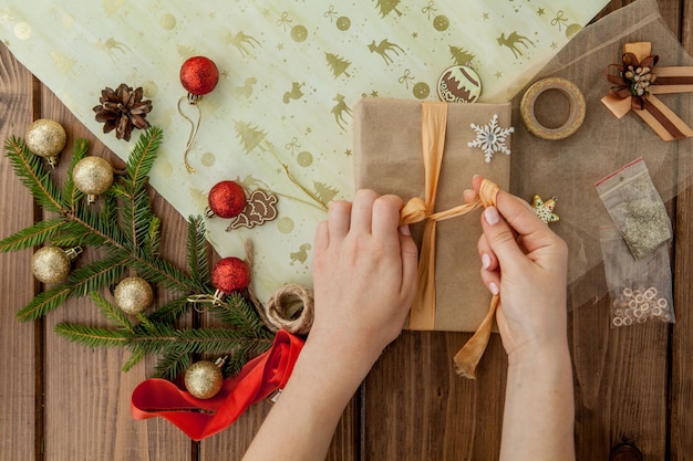 Die Hände der Frau, die oben Weihnachtsgeschenk, Abschluss einwickeln. Unvorbereitete Weihnachtsgeschenke auf hölzernem mit Dekorelementen und Einzelteilen, Draufsicht. Weihnachten oder Neujahr DIY Verpackung.