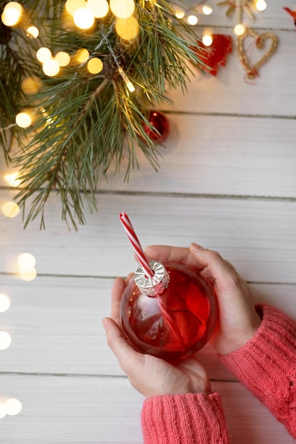 Die Hände der Frau, die rote Weihnachtsglaskugel mit einem Getränk auf weißem Tisch halten Authentisches Weihnachtsfoto