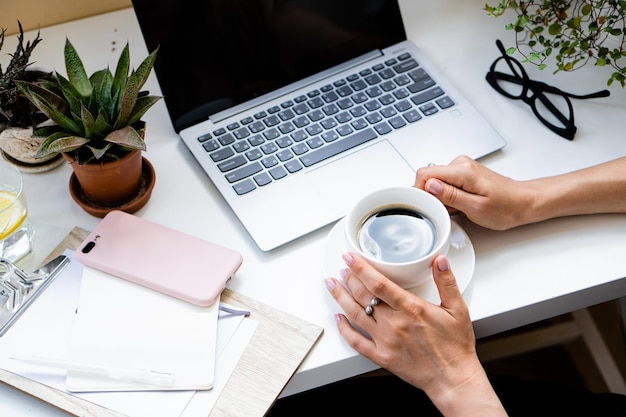 Die Hände der Frau halten eine Tasse Kaffee in einem gemütlichen Büro mit Laptop und grünen Pflanzen