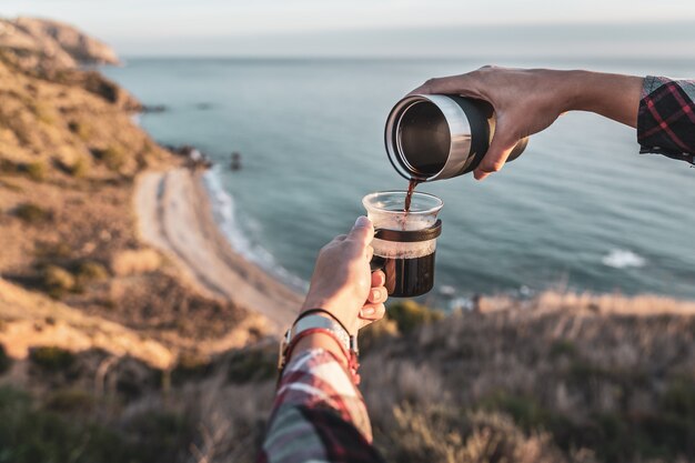 Die Hände der Frauen, die Kaffee in ihrer Schale mit der Küste im Hintergrund zubereiten. Konzept der Erkundung und Abenteuer