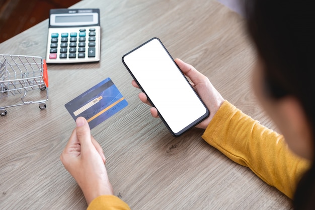 Die Hände der Frauen im gelben Hemd, das Handy mit leerem Bildschirm und Kreditkarte auf Schreibtisch hält und Handy für das on-line-Einkaufen, Internetbanking, on-line-Handel verwendet