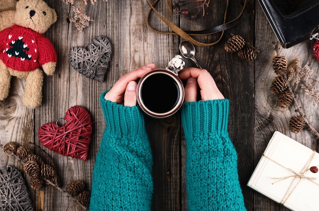 Die Hände der Frauen in einer grünen gestrickten Strickjacke, die einen roten keramischen Becher mit schwarzem Kaffee hält