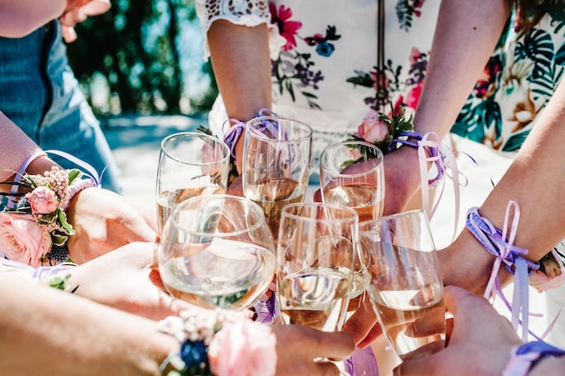 Die Hände der Mädchen mit Boutonnieres und Bändern halten und heben Gläser Champagner hoch Champagnerglas mit Blumen geschmückt Party im Stil Boho Jungfernabend Junggesellinnenabschiede Junggesellinnenabschied Nahaufnahme