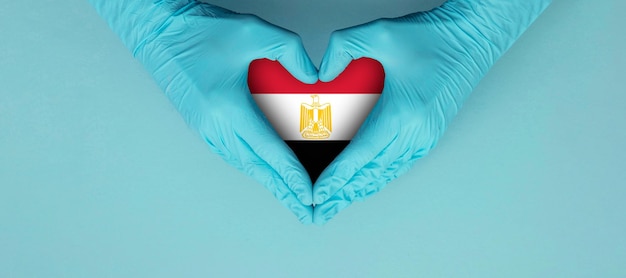 Die Hände des Arztes tragen blaue OP-Handschuhe und machen ein Hörformsymbol mit ägyptischer Flagge