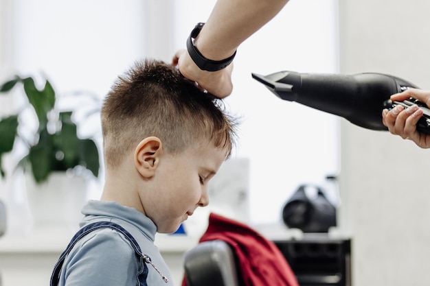 Die Hände des Friseurs trocknen die Haare des Jungen mit einem Fön Nahaufnahme der Frau, die die Haare des Jungen im Friseursalon pflegt Porträt des männlichen Kindes im Friseursalon, um seine Haare zu schneiden