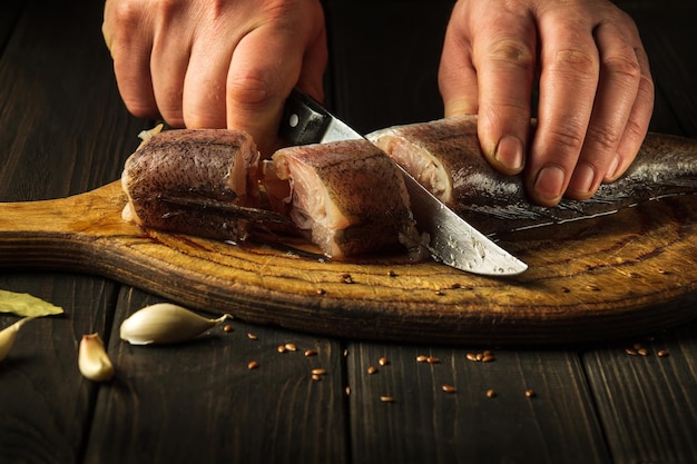 Die Hände des Küchenchefs mit einem Messer schneiden frischen Merluccius-Fisch auf einem Holzschneidebrett