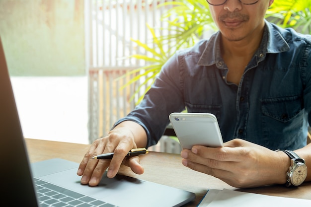 Die Hände des Mannes unter Verwendung des Handys beim Arbeiten mit Laptop auf hölzernem Schreibtisch