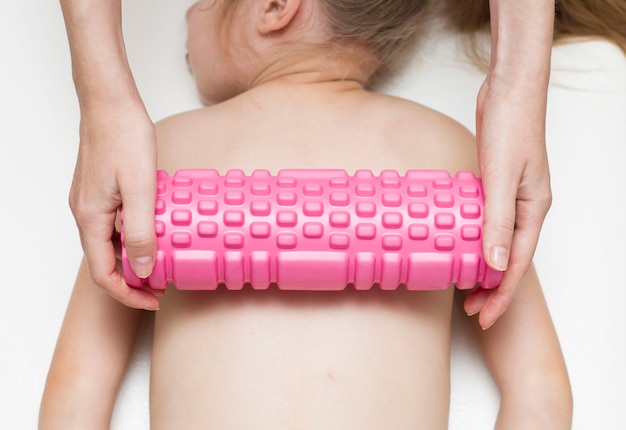 Die Hände des Masseurs machen eine Rückenmassage mit einer rosa Rolle für ein Mädchen im Grundschulalter. Wirbelsäule. Rückenschmerzen bei schulpflichtigen Kindern.