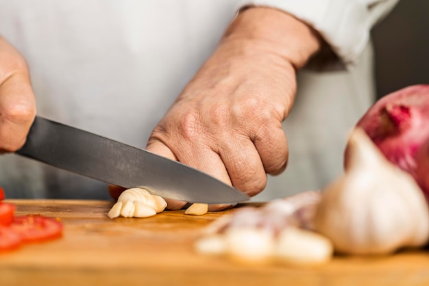 Die Hände einer erwachsenen Frau, die einen Knoblauch mit einem Küchenmesser auf einem Schneidebrett für die Lasagnezubereitung schneidet