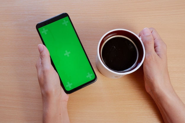 Die Hände einer jungen Frau mit einem Greenscreen-Telefon und einer Tasse Kaffee am Tisch