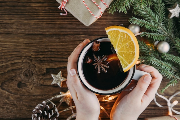 Die Hände halten eine Tasse mit Weihnachts- und Neujahrsgetränk, heißem Wein, Glühwein, Punsch oder Tee auf einem Holztisch neben einem grünen Weihnachtsbaum und einer Girlande. Platz für Text.