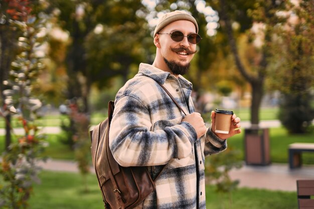 Die halbe Länge des glücklichen Gesichts des Kerls, der im Freien im Park herumläuft, hält Kaffee zum Mitnehmen, während Sie sich entspannen