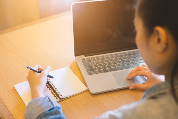 Die Hand der Frau, die Laptop verwendet und in Notizblock schreibt.