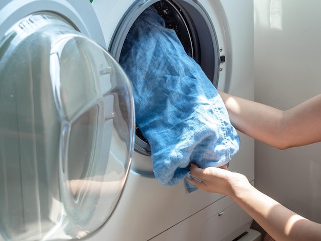 Die Hand der Frau, die schmutziges blaues Leinenhemd in die Waschmaschine legt