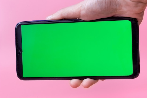 Die Hand der Frau hält das Smartphone in einer horizontalen Position mit einem grünen Bildschirm auf einem rosa Hintergrund. Chroma-Key. Attrappe, Lehrmodell, Simulation