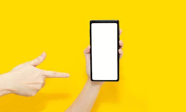 Die Hand der Frau hält ein Smartphone und zeigt mit dem Zeigefinger auf ein Smartphone mit weißem Bildschirm auf gelbem Hintergrund für Werbung und Promotion.