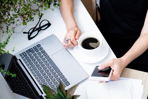 Die Hand der Frau hält eine Tasse Kaffee und ein Smartphone in einem gemütlichen Büro mit Laptop und grünen Pflanzen